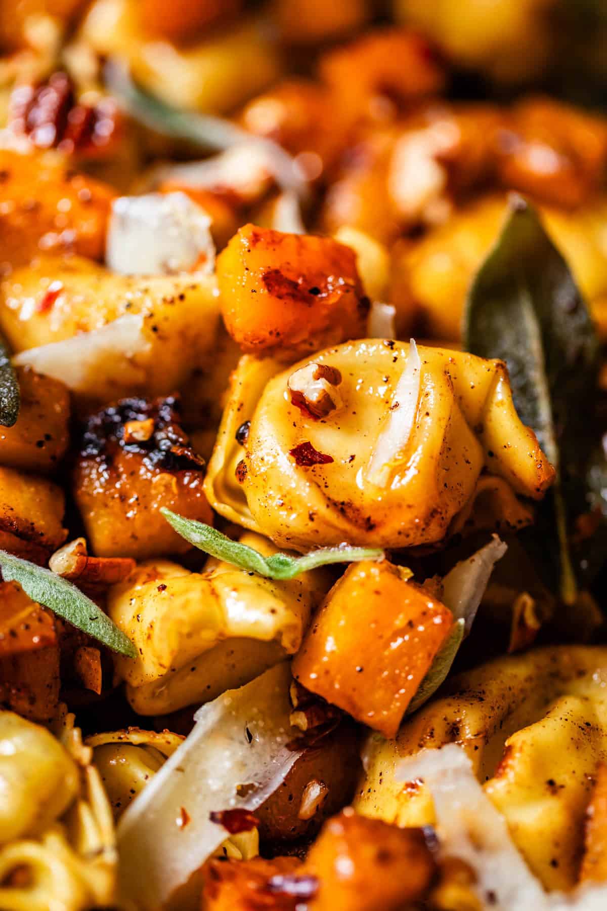 Close-up of tortellini pasta mixed with roasted butternut squash, fresh sage leaves, shaved parmesan cheese, and red pepper flakes. The dish appears flavorful and seasoned.