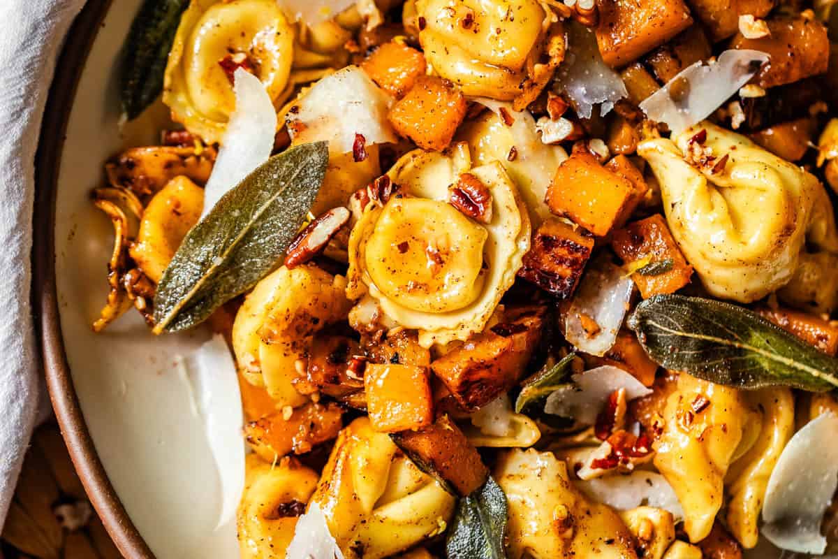 A close-up of a dish featuring cheese tortellini, roasted butternut squash, crispy sage leaves, pecans, and shaved Parmesan on a plate. The ingredients are golden and garnished with spices.