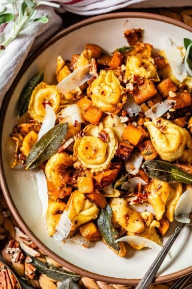 A plate of cheese tortellini mixed with roasted butternut squash, crispy sage leaves, shaved parmesan, pecans, and spices, served with a fork. The dish has a golden, autumn-inspired appearance.