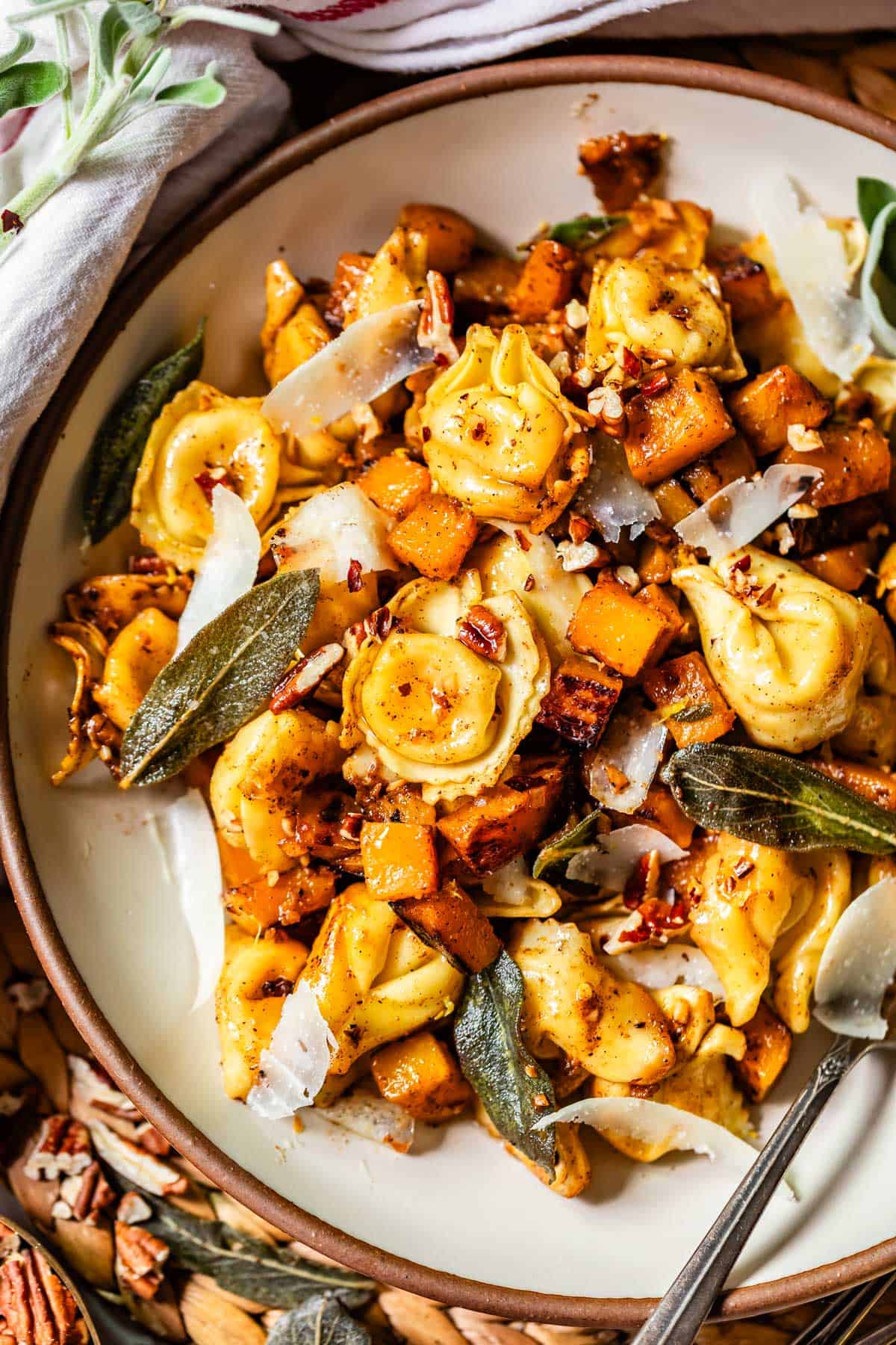 A plate of cheese tortellini mixed with roasted butternut squash, crispy sage leaves, shaved parmesan, pecans, and spices, served with a fork. The dish has a golden, autumn-inspired appearance.