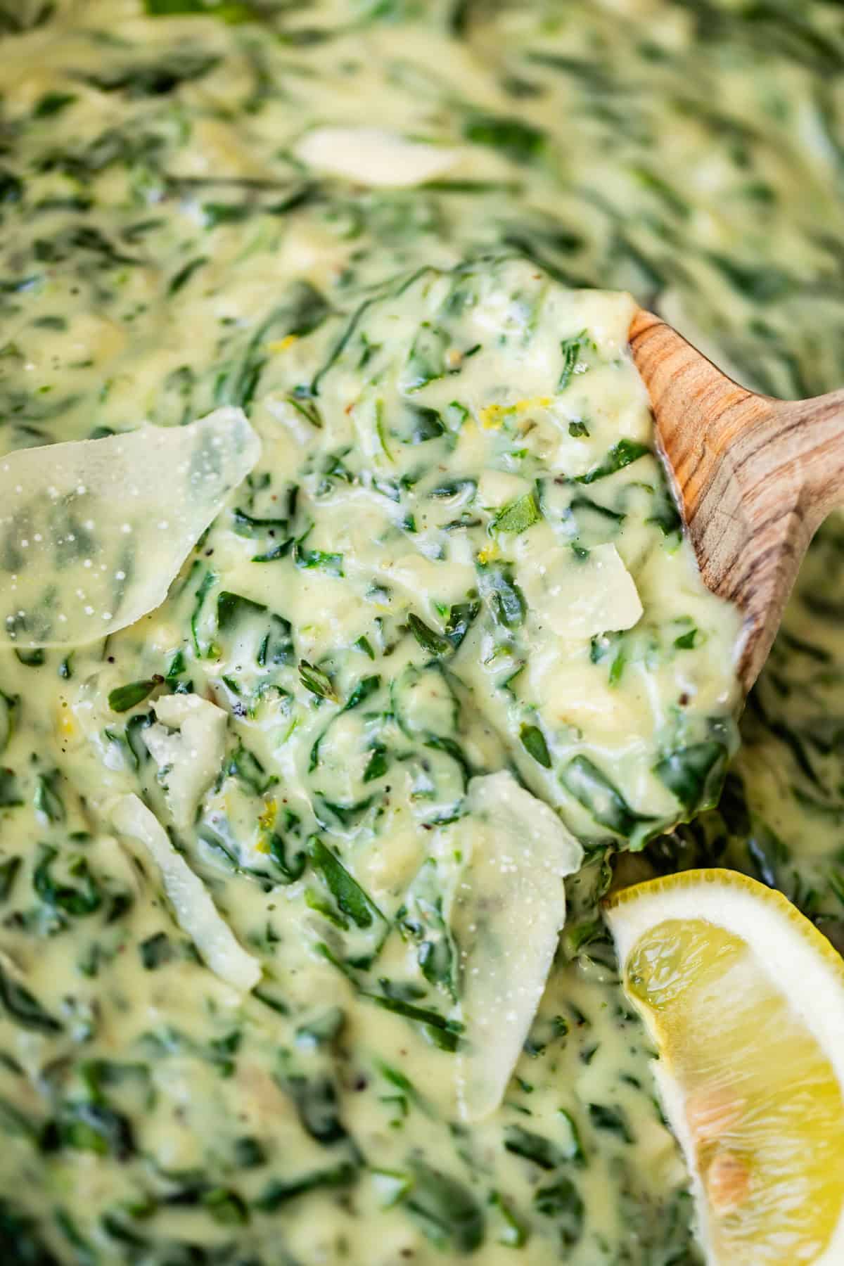 Close-up of creamy spinach dip with visible cheese shavings, held up by a wooden spoon. A wedge of lemon is positioned on the side of the dish.