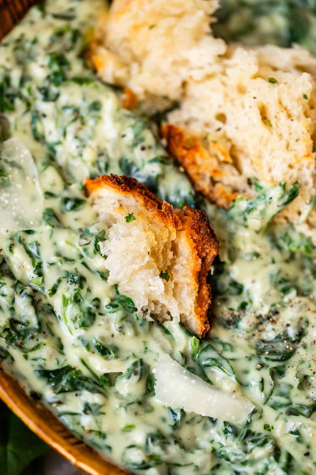Close-up of creamy spinach dip with visible herbs and cheese, topped with pieces of crusty bread for dipping.
