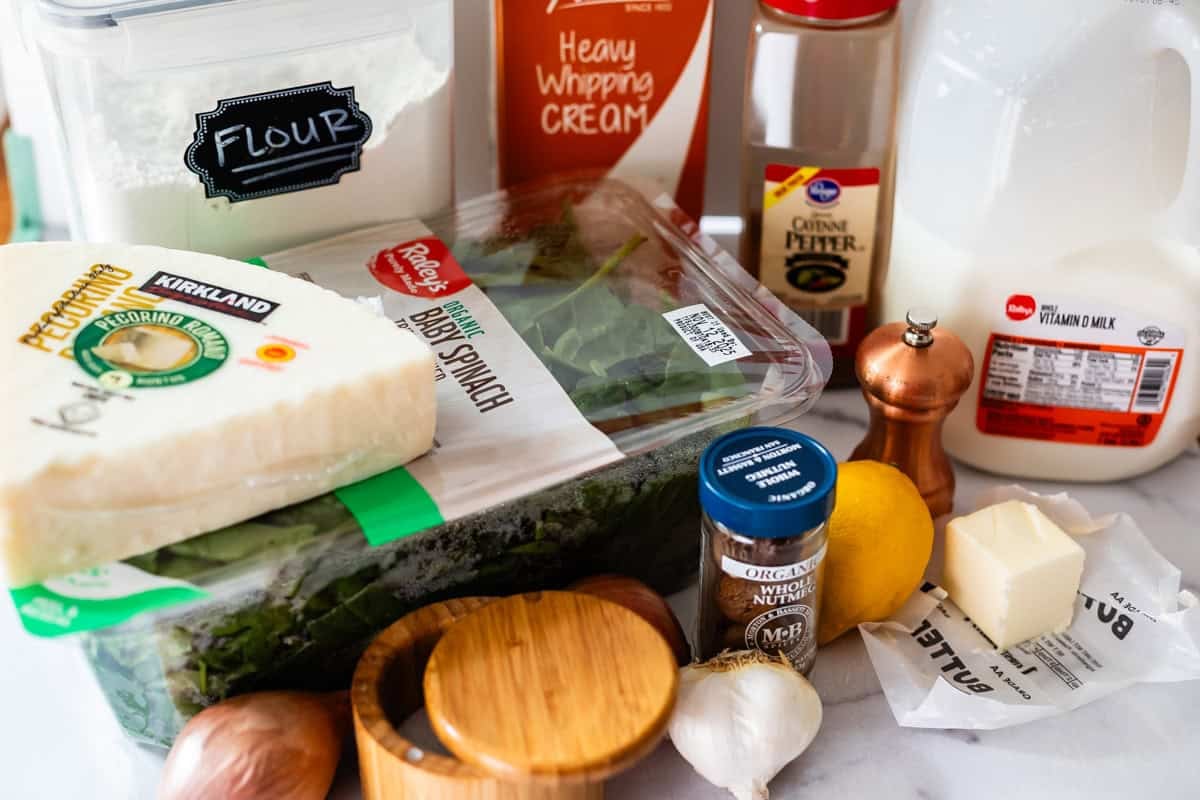 A collection of cooking ingredients on a countertop, including flour, heavy whipping cream, milk, pecorino cheese, baby spinach, black pepper, a lemon, garlic, nutmeg, butter, and a wooden salt container.