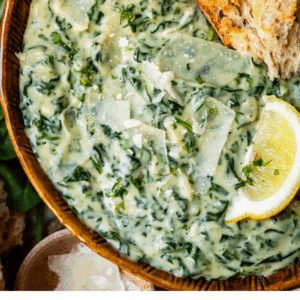 A bowl of creamy spinach topped with Parmesan shavings and a lemon wedge, served with a piece of crusty bread. Text at the bottom reads Creamed Spinach Youll Actually Like - The Food Charlatan.