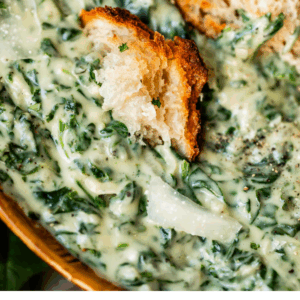 Close-up of creamy creamed spinach topped with a couple of pieces of rustic bread. The dish is rich and green, with visible spinach and creamy sauce. Text below reads: CREAMED SPINACH YOULL ACTUALLY LIKE.