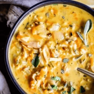 A close-up of a bowl of creamy chicken and wild rice soup, garnished with fresh herbs, with a spoon resting inside. The soup is thick, yellowish, and filled with visible pieces of chicken and vegetables.