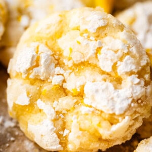 Close-up of a lemon crinkle cookie covered in powdered sugar, with a cracked surface and a bright yellow color. Blurred lemon slices and more cookies are visible in the background.