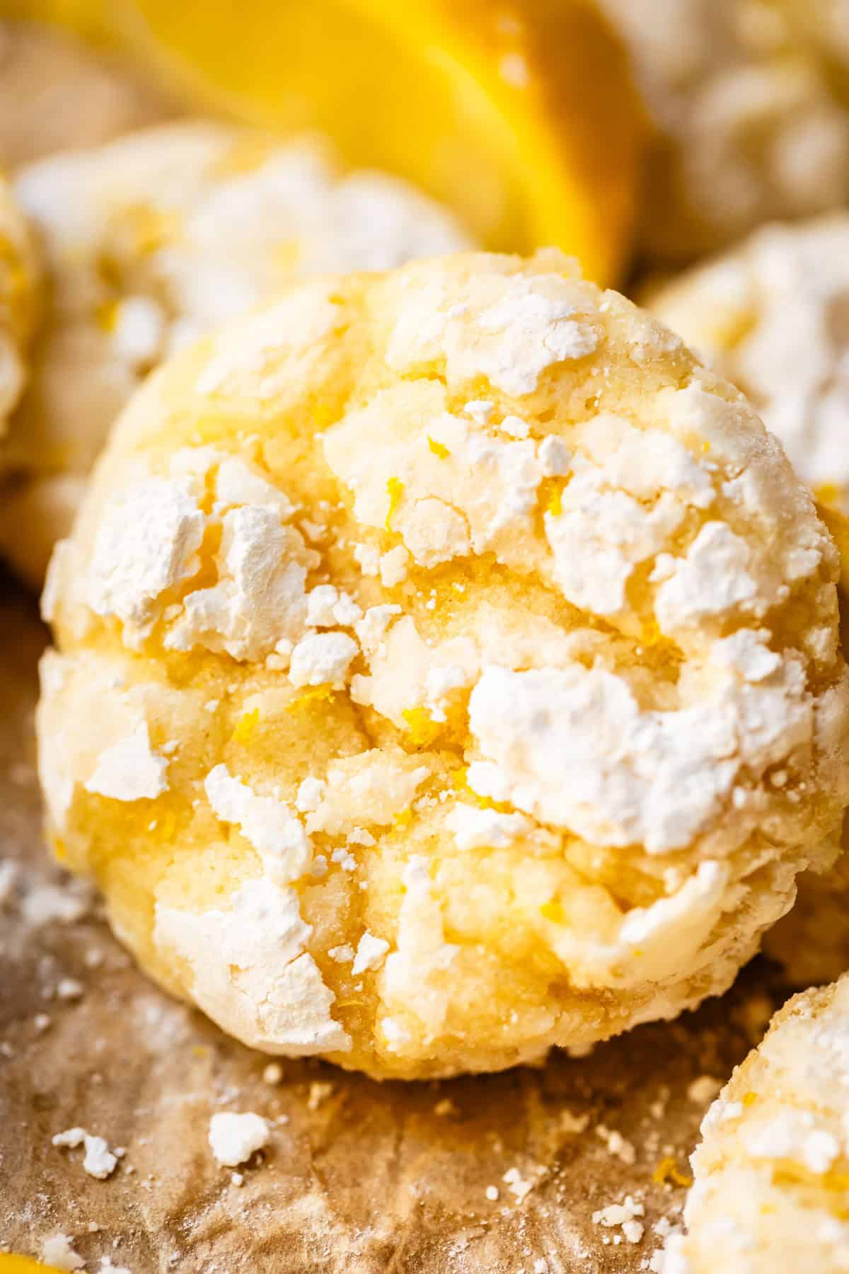 Close-up of a lemon crinkle cookie covered in powdered sugar, with a cracked surface and a bright yellow color. Blurred lemon slices and more cookies are visible in the background.