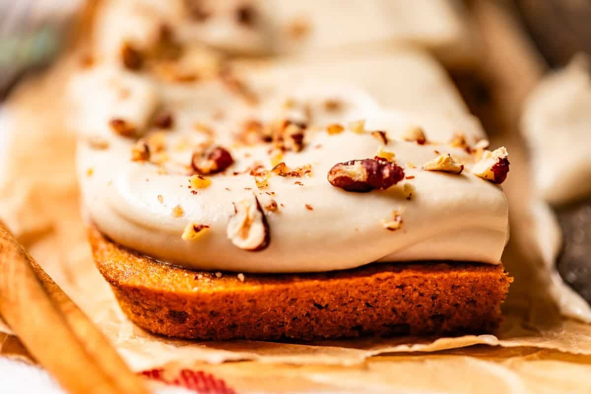 A close-up of a frosted bar dessert topped with chopped nuts on parchment paper. The thick, creamy frosting is spread over a golden-brown base, with nut pieces sprinkled on top.