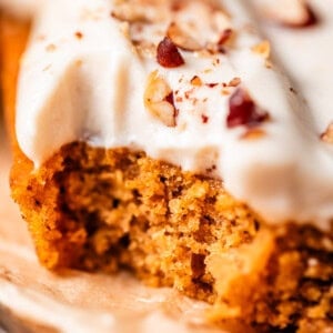 A close-up of a slice of moist, spiced cake topped with creamy white frosting and sprinkled with chopped nuts, resting on a piece of parchment paper.