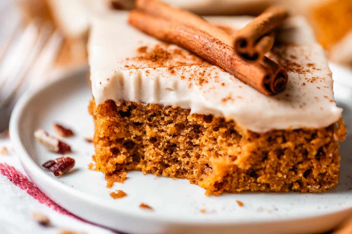 A close-up of a slice of spiced cake topped with white frosting and two cinnamon sticks on a white plate, with crumbs and pecans scattered nearby.