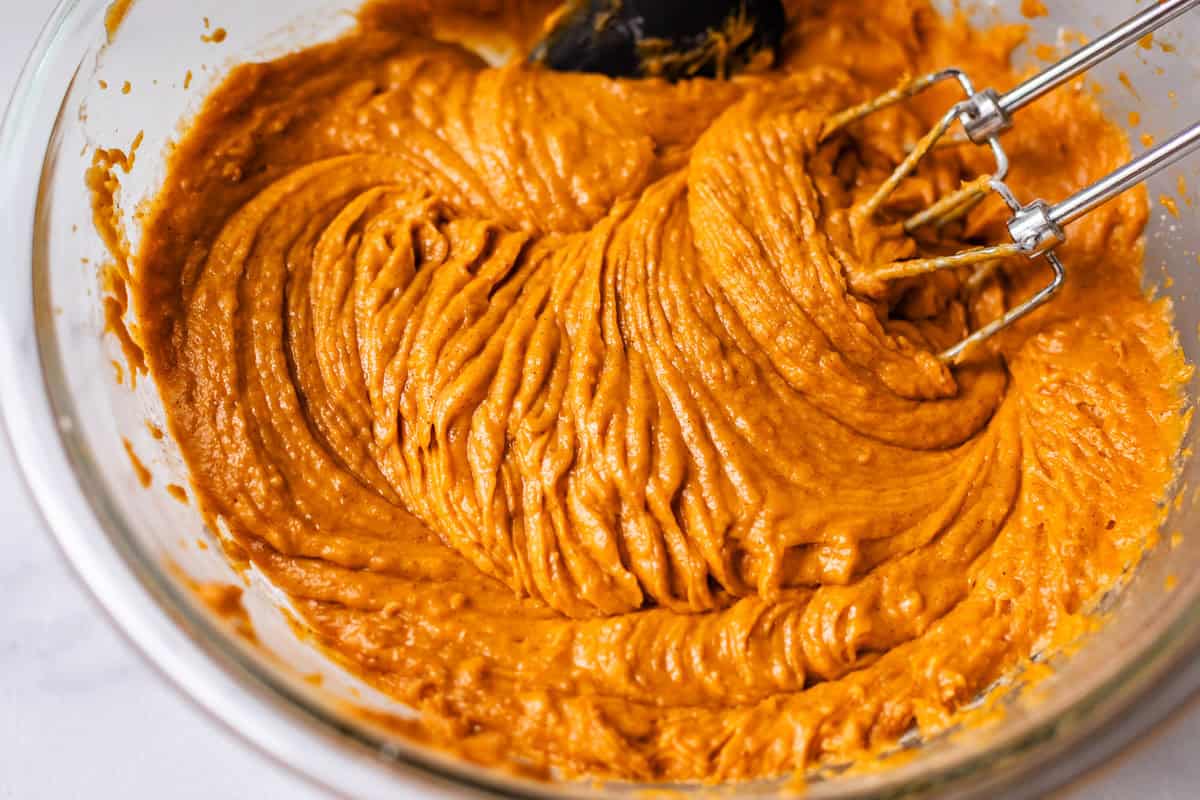 Close-up of a glass mixing bowl filled with thick, orange pumpkin batter being mixed with an electric hand mixer.