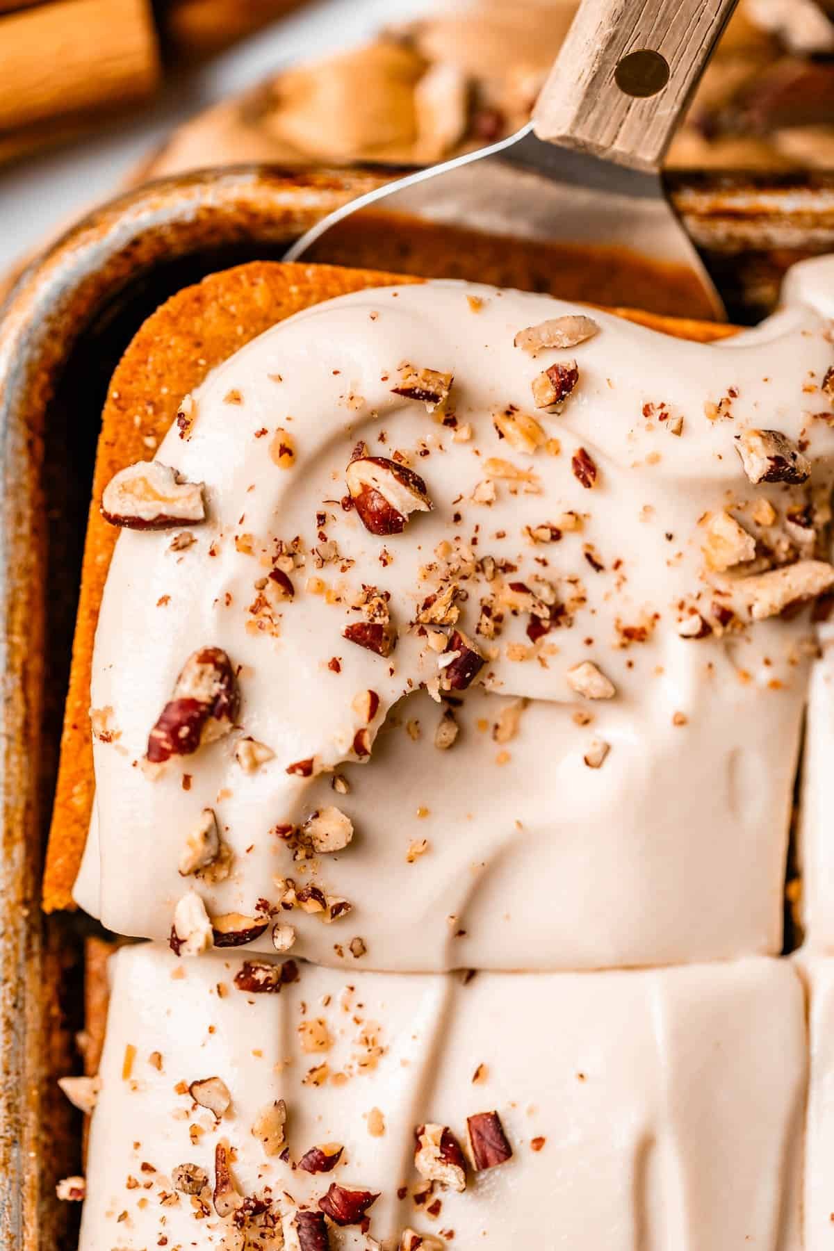 HERO A spatula lifts a square piece of pumpkin sheet cake topped with creamy white frosting and sprinkled with chopped pecans from a baking pan.