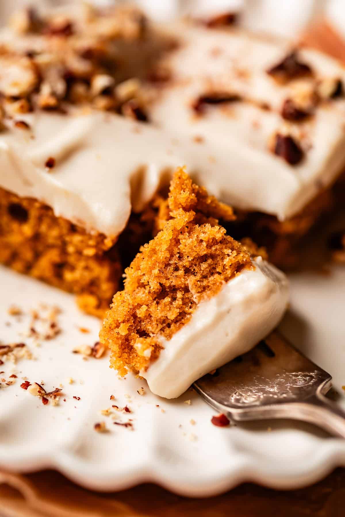 A close-up of a moist slice of carrot cake topped with creamy frosting and chopped nuts, resting on a decorative plate with a fork holding a bite-sized piece.
