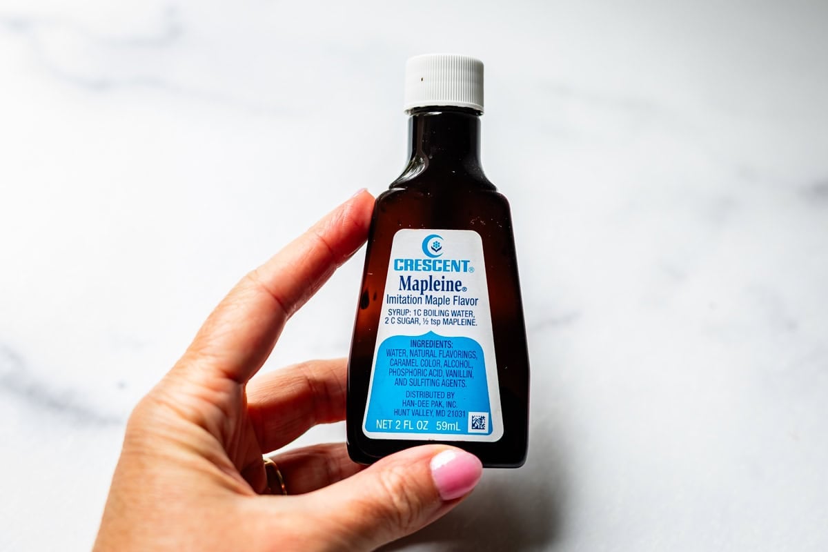 A hand holding a small dark brown bottle of Crescent Mapleine imitation maple flavoring with a white cap and blue-and-white label, against a white background.