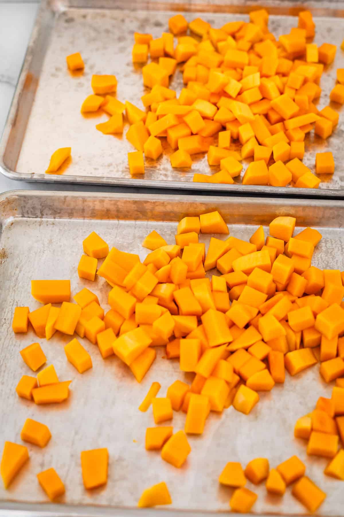 Two metal baking sheets topped with evenly spaced, small cubes of bright orange butternut squash, ready to be roasted.
