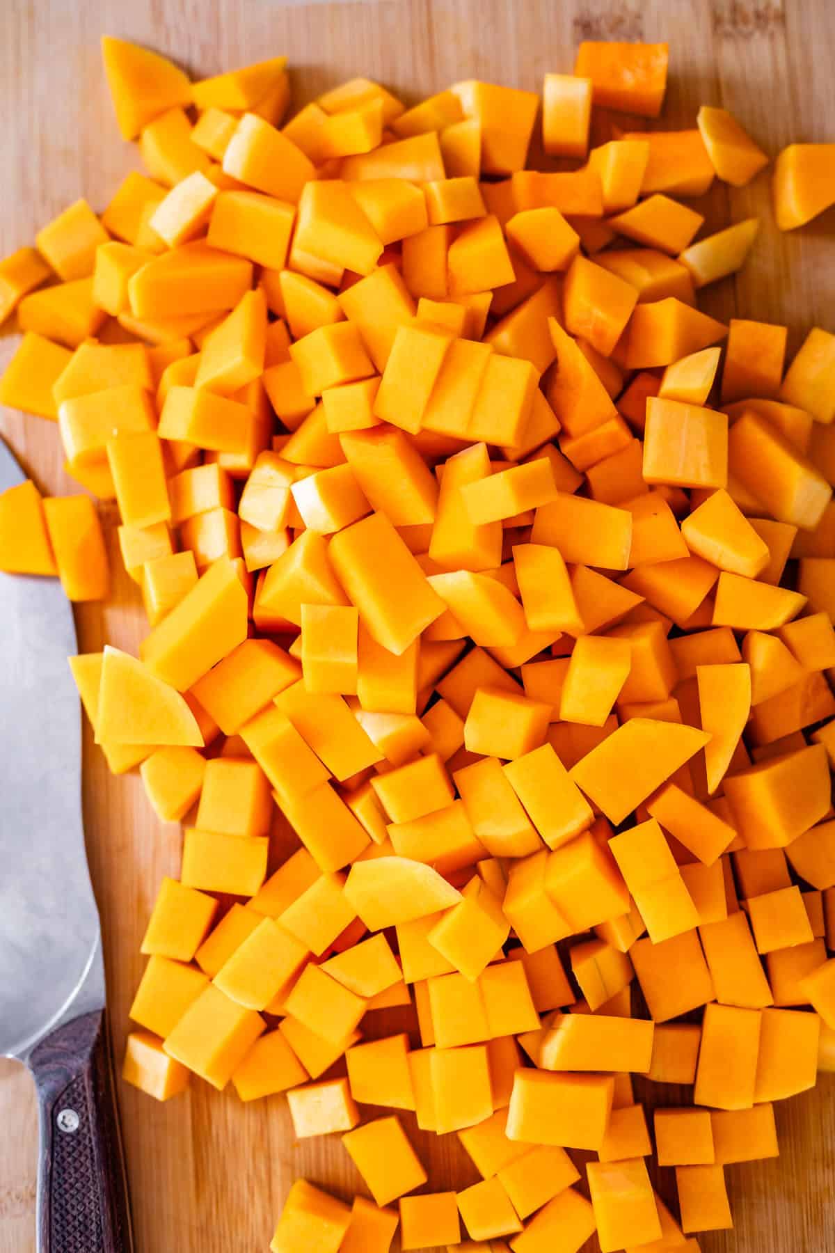 Diced butternut squash pieces on a wooden cutting board next to a chef’s knife with a dark handle.