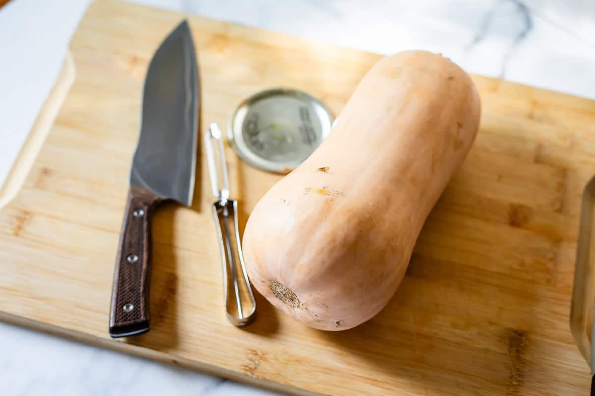 A whole butternut squash, a chef’s knife, a vegetable peeler, and a metal jar lid rest on a wooden cutting board on a light-colored surface.