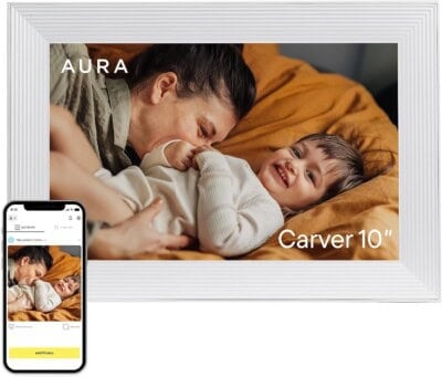 A white digital photo frame displays a smiling child lying on a bed with an adult; a smartphone nearby shows the same photo and an app interface. Text on frame reads AURA Carver 10”.