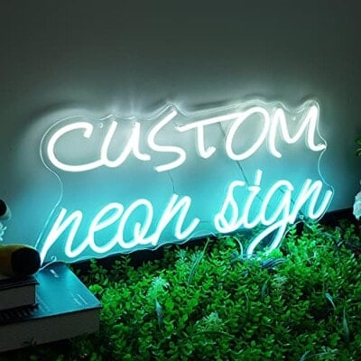 A glowing neon sign with the words custom neon sign in white and blue, placed above green artificial grass and next to stacked books.