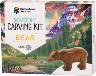 A box labeled Soapstone Carving Kit Bear by Studiostone Creative, featuring a carved bear figurine and a colorful illustrated mountain landscape in the background. Suitable for ages 8 to 99.
