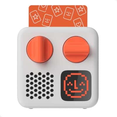 A small white audio device with two large orange knobs, a speaker, a digital screen displaying a smiling pixelated face, and an orange card with playful designs inserted at the top.