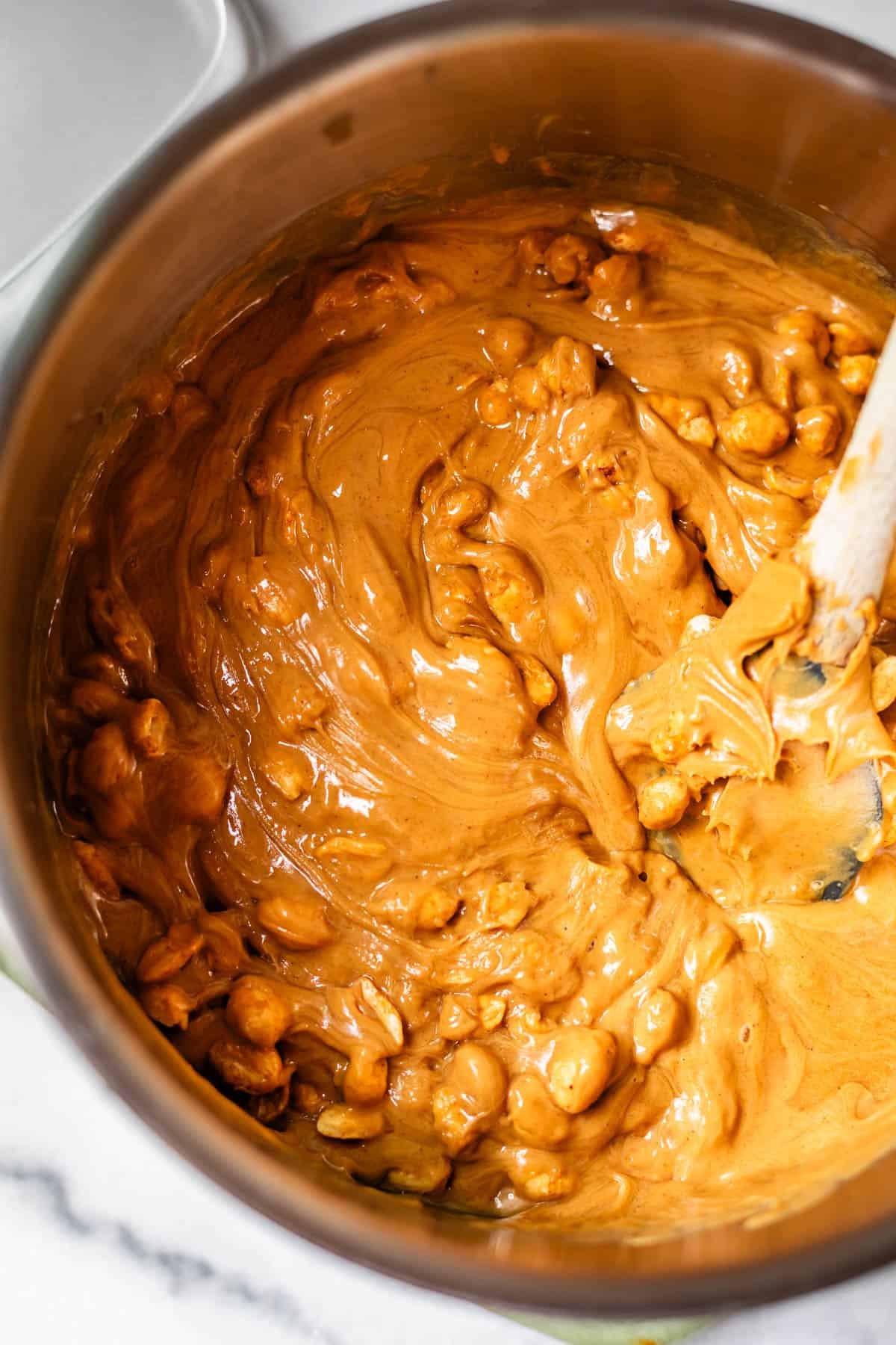 A saucepan filled with a thick, creamy, caramel-colored mixture and peanuts being stirred with a white spatula on a marble countertop.