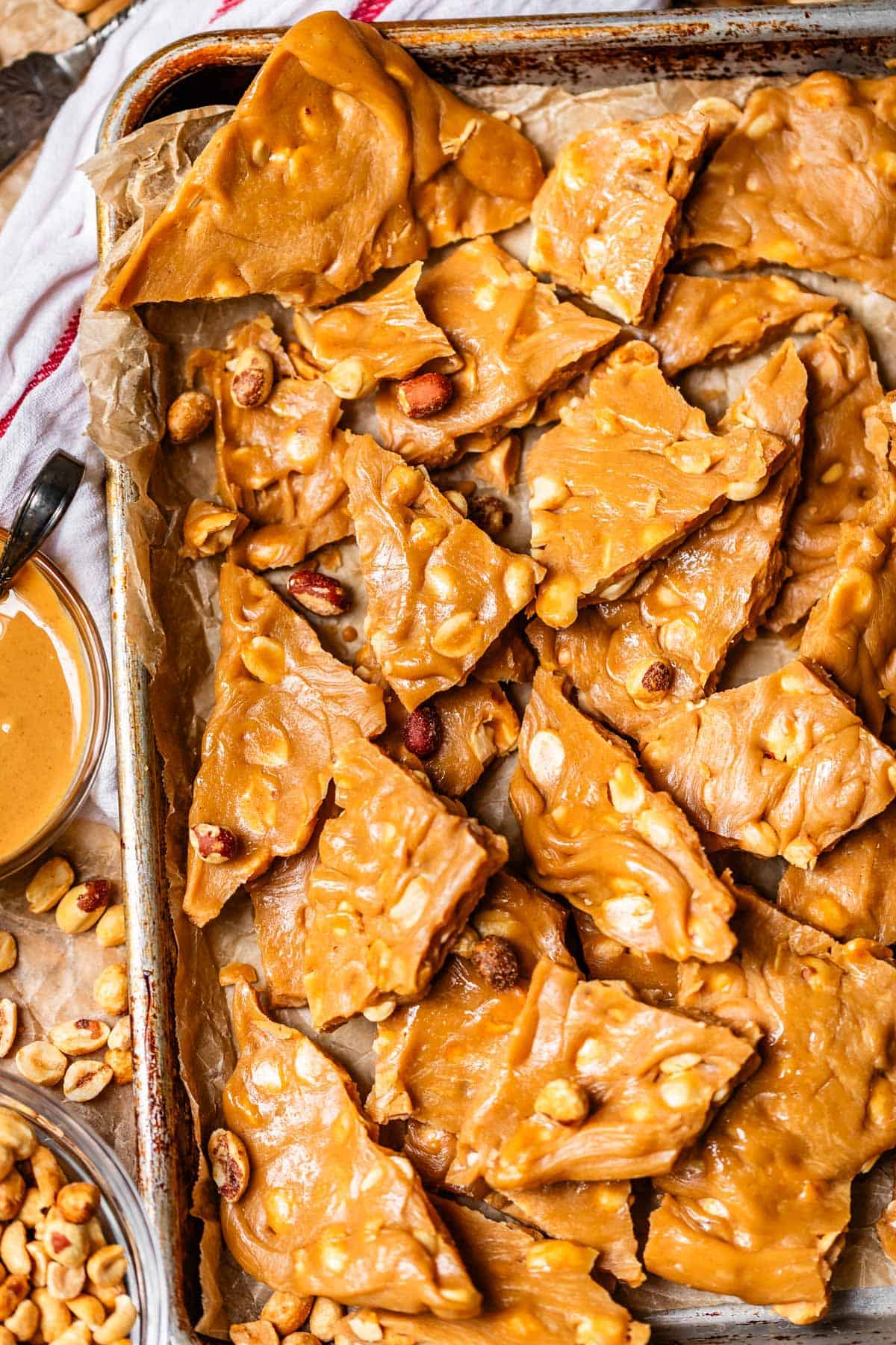 A baking tray lined with parchment paper holds broken pieces of golden brown peanut brittle with visible peanuts. A small bowl of peanut butter and scattered peanuts are beside the tray.