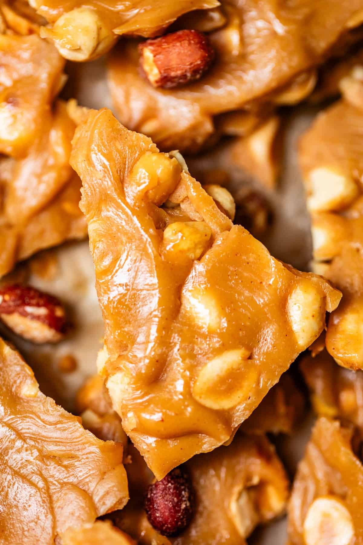 Close-up of golden peanut brittle pieces, showing a glossy, caramelized surface with embedded roasted peanuts. The texture appears crunchy and slightly uneven, highlighting the nutty, sugary treat.