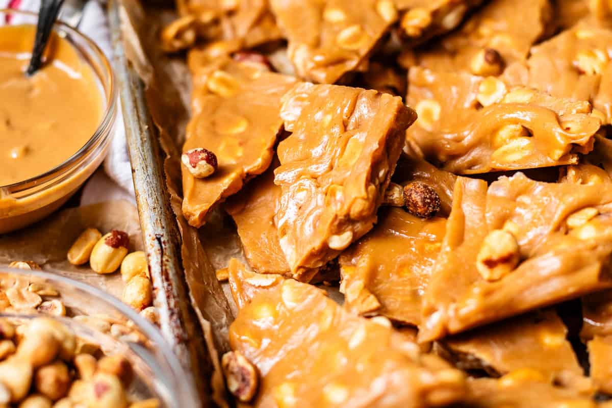 Close-up of golden, crunchy peanut brittle pieces stacked on a tray, with visible whole peanuts embedded in the caramelized candy. A glass bowl of peanut butter and extra peanuts are seen nearby.