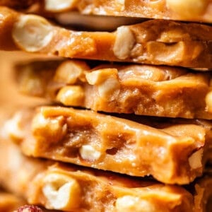 Close-up of stacked pieces of peanut brittle showing a golden, caramelized candy filled with whole and chopped peanuts. The surface looks glossy and textured with visible nut pieces throughout.