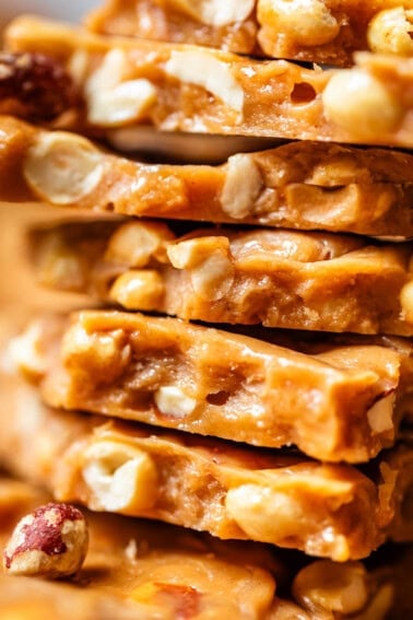 Close-up of stacked pieces of peanut brittle showing a golden, caramelized candy filled with whole and chopped peanuts. The surface looks glossy and textured with visible nut pieces throughout.