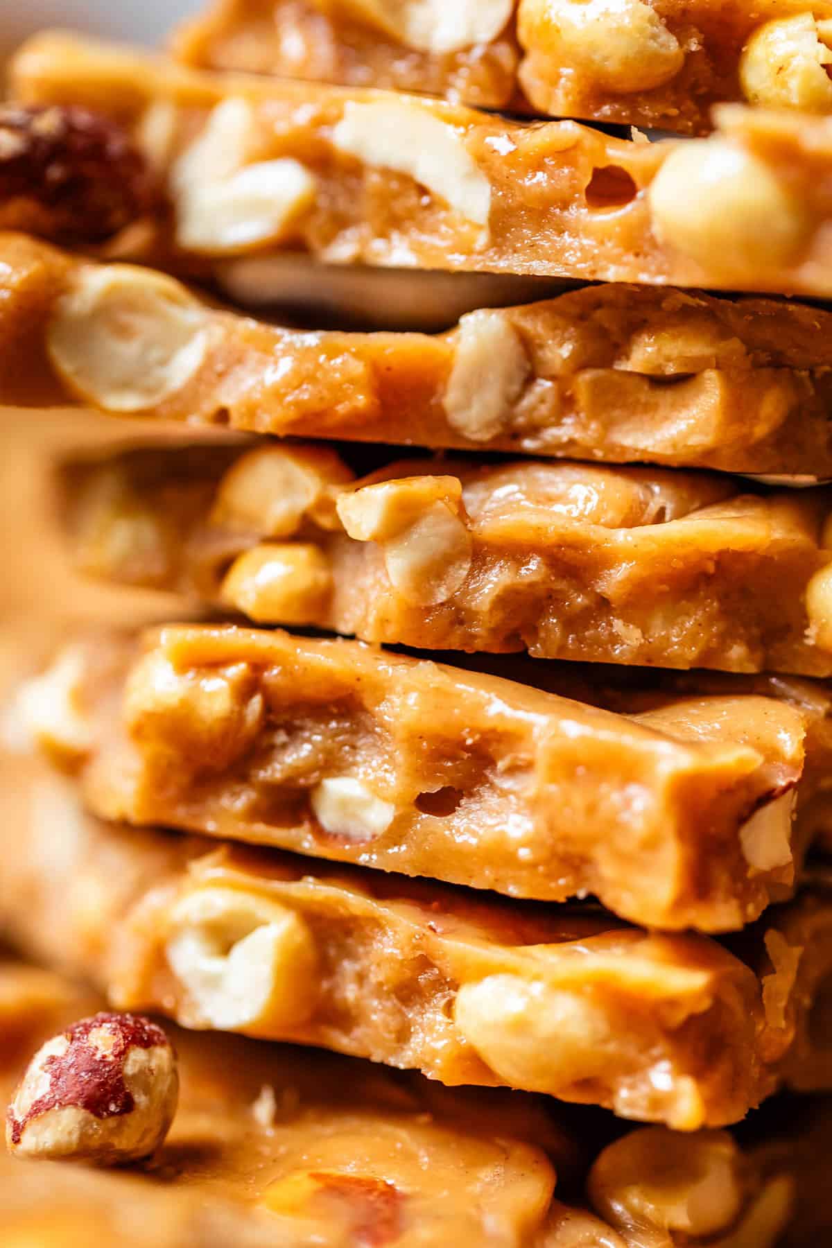 Close-up of stacked pieces of peanut brittle showing a golden, caramelized candy filled with whole and chopped peanuts. The surface looks glossy and textured with visible nut pieces throughout.
