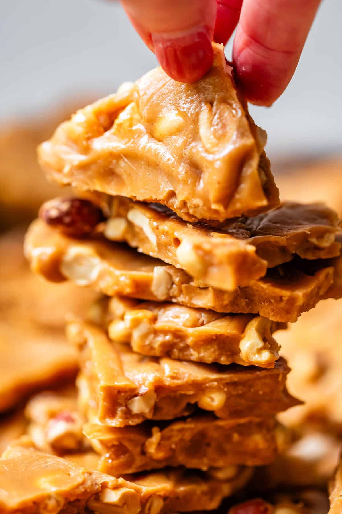 A close-up of a hand picking up a piece of golden, nut-filled peanut brittle from the top of a stacked pile of brittle pieces. The candy is glossy and crunchy, with visible whole peanuts inside.