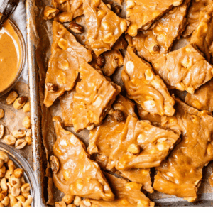 A baking sheet lined with parchment paper holds broken pieces of homemade peanut brittle, surrounded by peanuts and a bowl of peanut butter. Text at the bottom reads: Simple Homemade Peanut Brittle.