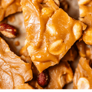 Close-up of golden, shiny peanut brittle pieces with visible peanuts, arranged closely together. Text at the bottom reads: Old-Fashioned Peanut Brittle - The Food Charlatan.