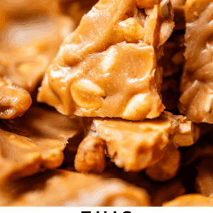 Close-up of golden, crunchy peanut brittle pieces stacked together, showing peanuts embedded in the caramelized candy. Text at the bottom reads: This Peanut Brittle is legit. The Food Charlatan.