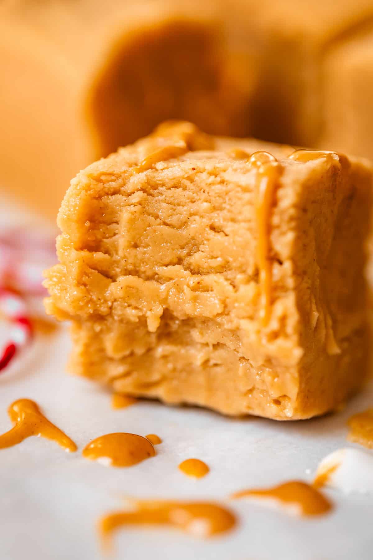 A close-up of a thick, creamy square of peanut butter fudge with a drizzle of melted peanut butter on top, sitting on parchment paper with extra drips around it.
