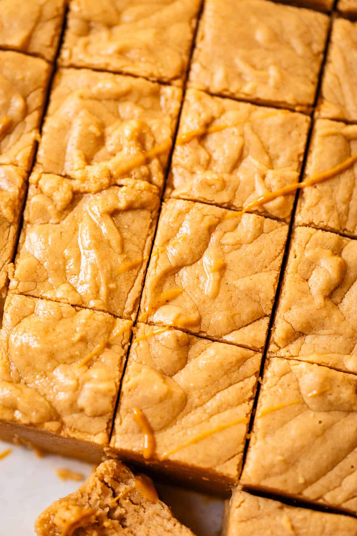 Close-up of soft, golden peanut butter fudge cut into squares, with a smooth, slightly textured surface and a light drizzle of peanut butter on top. One bar at the bottom is slightly pulled out.