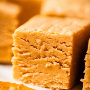 Close-up of a thick, creamy square of peanut butter fudge, showing its smooth and slightly crumbly texture. Other pieces of fudge are visible in the background.