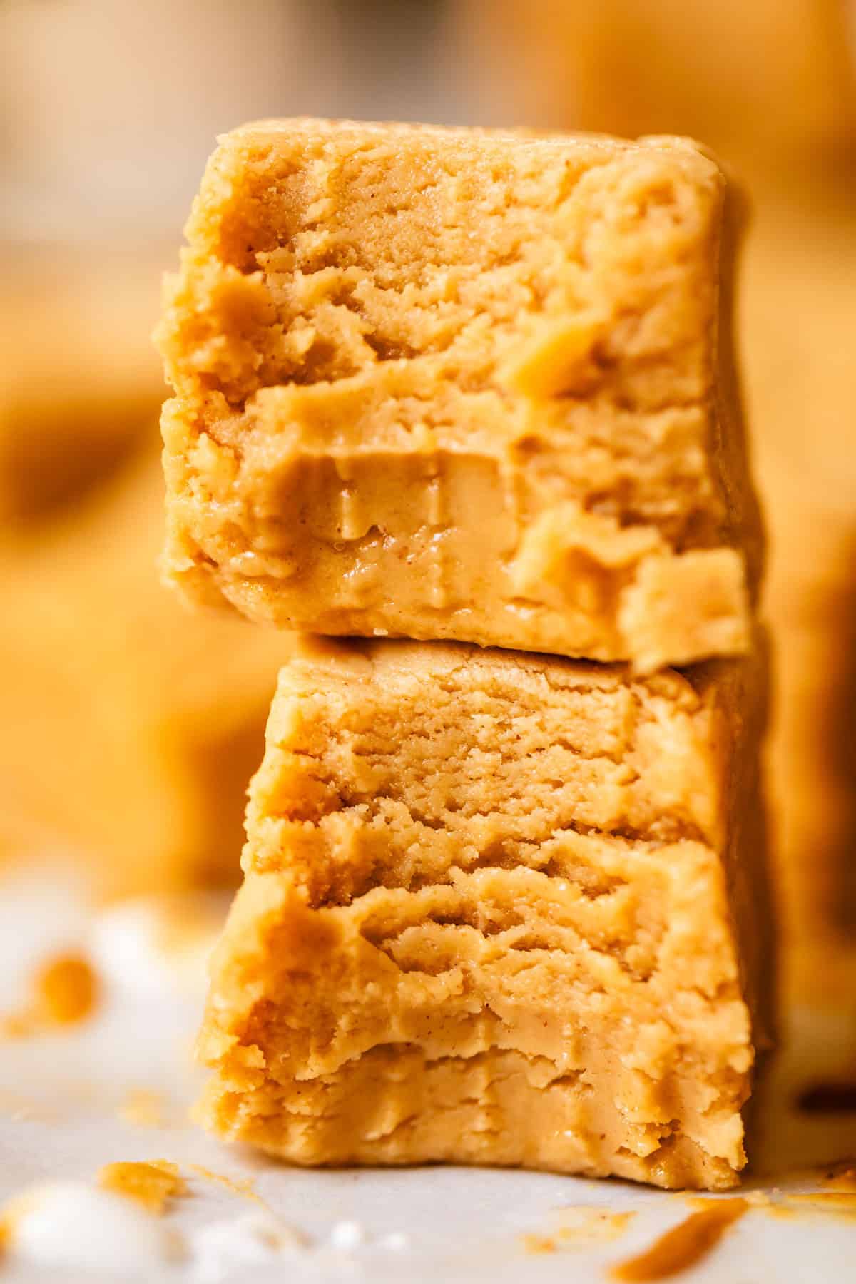 Two thick, creamy, golden-brown squares of peanut butter fudge are stacked on top of each other, showing a soft and smooth texture with some bite marks and crumbly edges.
