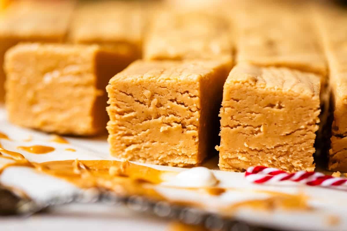 Close-up of creamy, square pieces of peanut butter fudge on a white surface, with a drizzle of peanut butter and a small piece of red and white striped candy in the foreground.