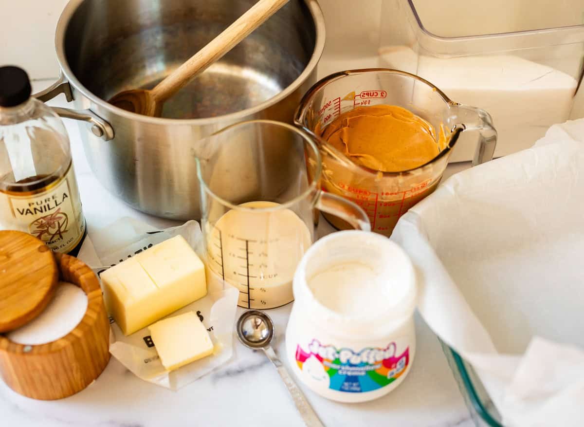Ingredients and tools for making fudge: a saucepan, wooden spoon, vanilla extract, butter, sugar, measuring cups with cream and peanut butter, marshmallow creme, and a parchment-lined baking dish on a countertop.