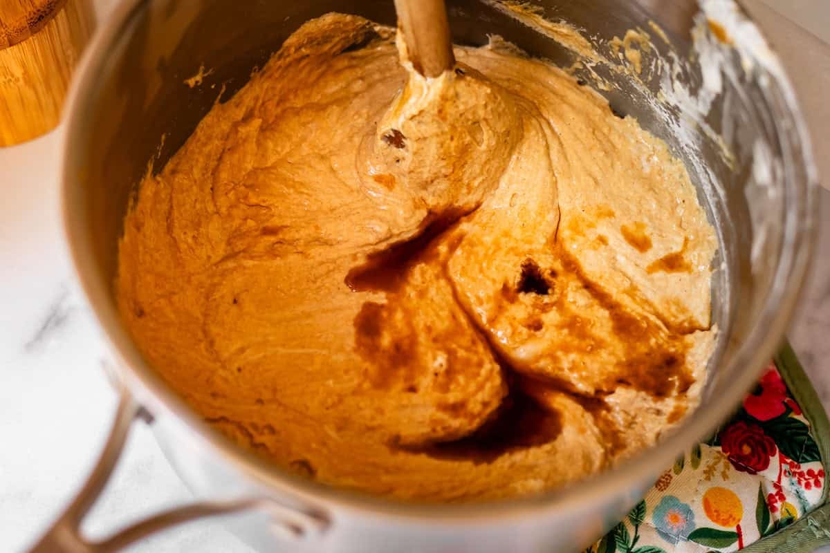 A metal mixing bowl filled with thick, light brown batter being stirred with a wooden spoon, with a splash of vanilla extract on top and a colorful floral oven mitt nearby.