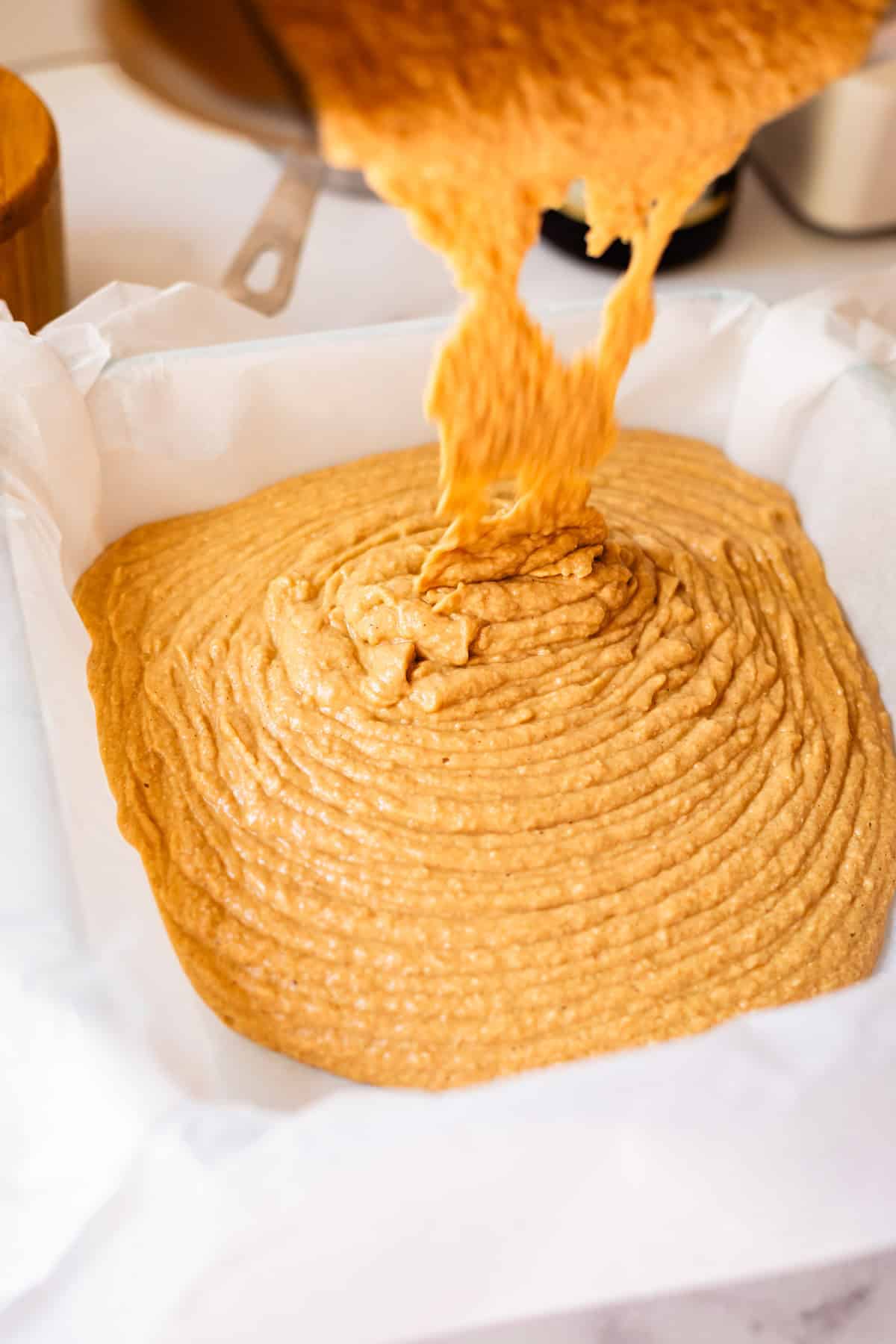 Thick, golden-brown fudge being poured into a parchment-lined baking pan, filling the pan with swirls and ridges.