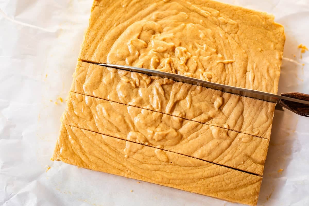 A close-up of a knife cutting through a large square block of peanut butter fudge on white parchment paper. The fudge has a creamy, swirled surface and is being sliced into rectangular pieces.