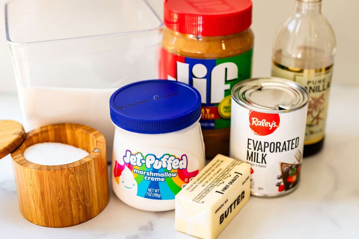 A collection of baking ingredients on a counter: a container of sugar, a wooden salt box, a jar of peanut butter, vanilla extract, Jet-Puffed marshmallow creme, a can of evaporated milk, and a stick of butter.