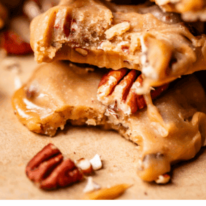 Close-up of creamy pecan pralines stacked on each other, showing their gooey texture and whole pecan pieces, with a few scattered pecans on a light surface. Text at the bottom reads Creamy Pecan Pralines.