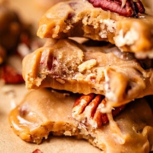 Close-up of stacked pecan pralines with a creamy caramel coating, showing a chewy texture and whole pecans. A few pralines and pecan pieces are scattered on a brown surface.