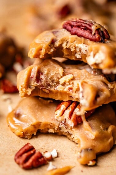 Close-up of stacked pecan pralines with a creamy caramel coating, showing a chewy texture and whole pecans. A few pralines and pecan pieces are scattered on a brown surface.